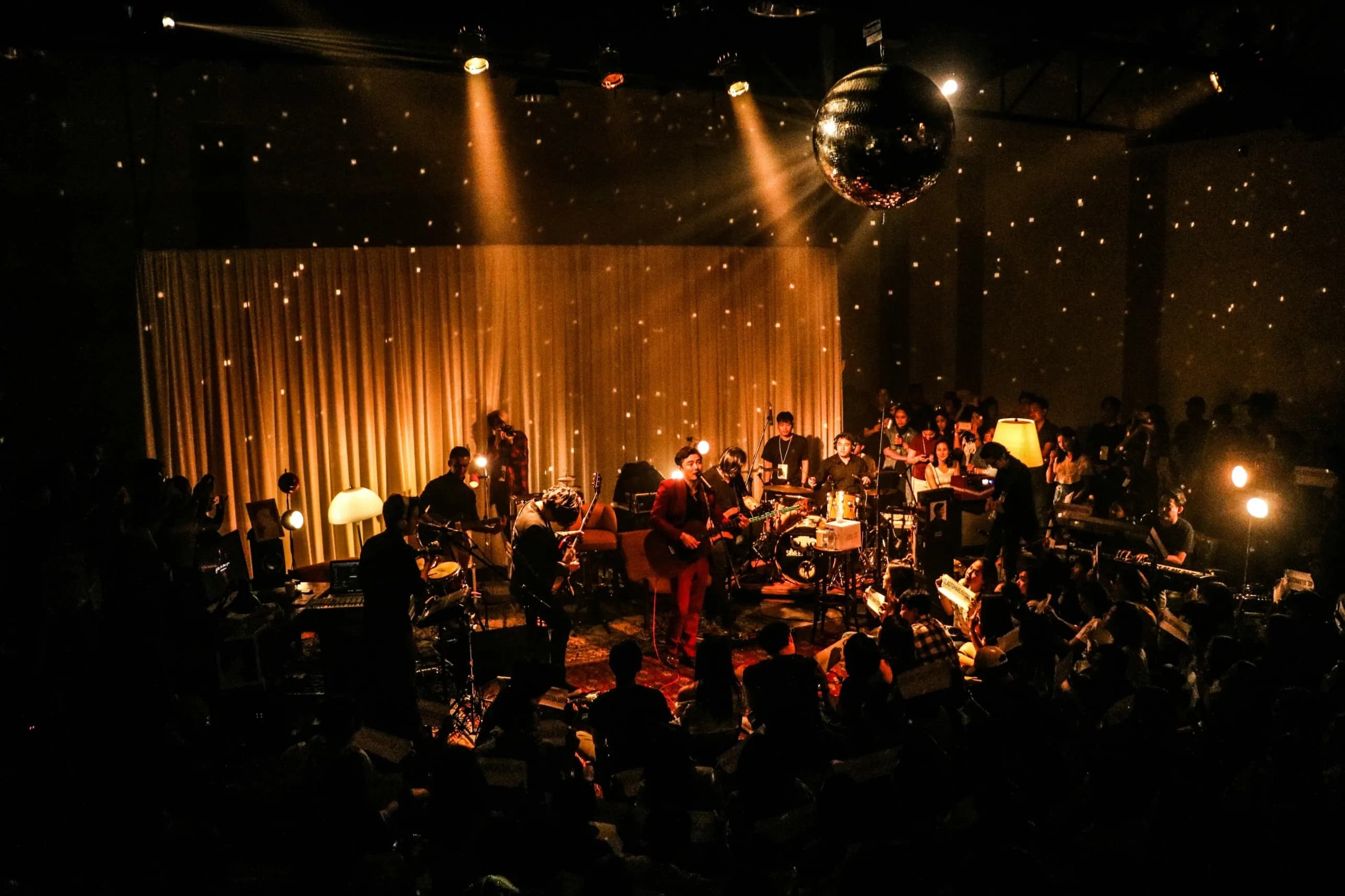 Un groupe joue devant un rideau doré constellé de lumières, au milieu d’un public assis tout près des musiciens.