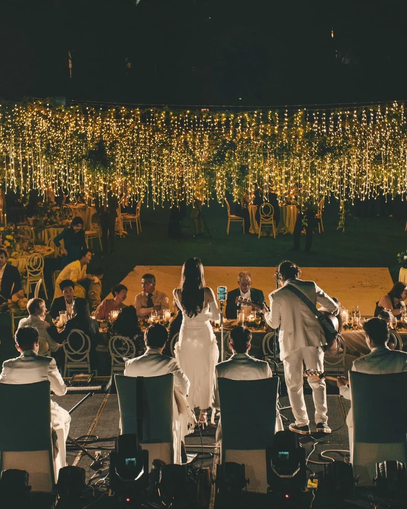Un groupe vêtu de blanc joue au plus près des tables d’un dîner en plein air, sous un ciel de guirlandes lumineuses.