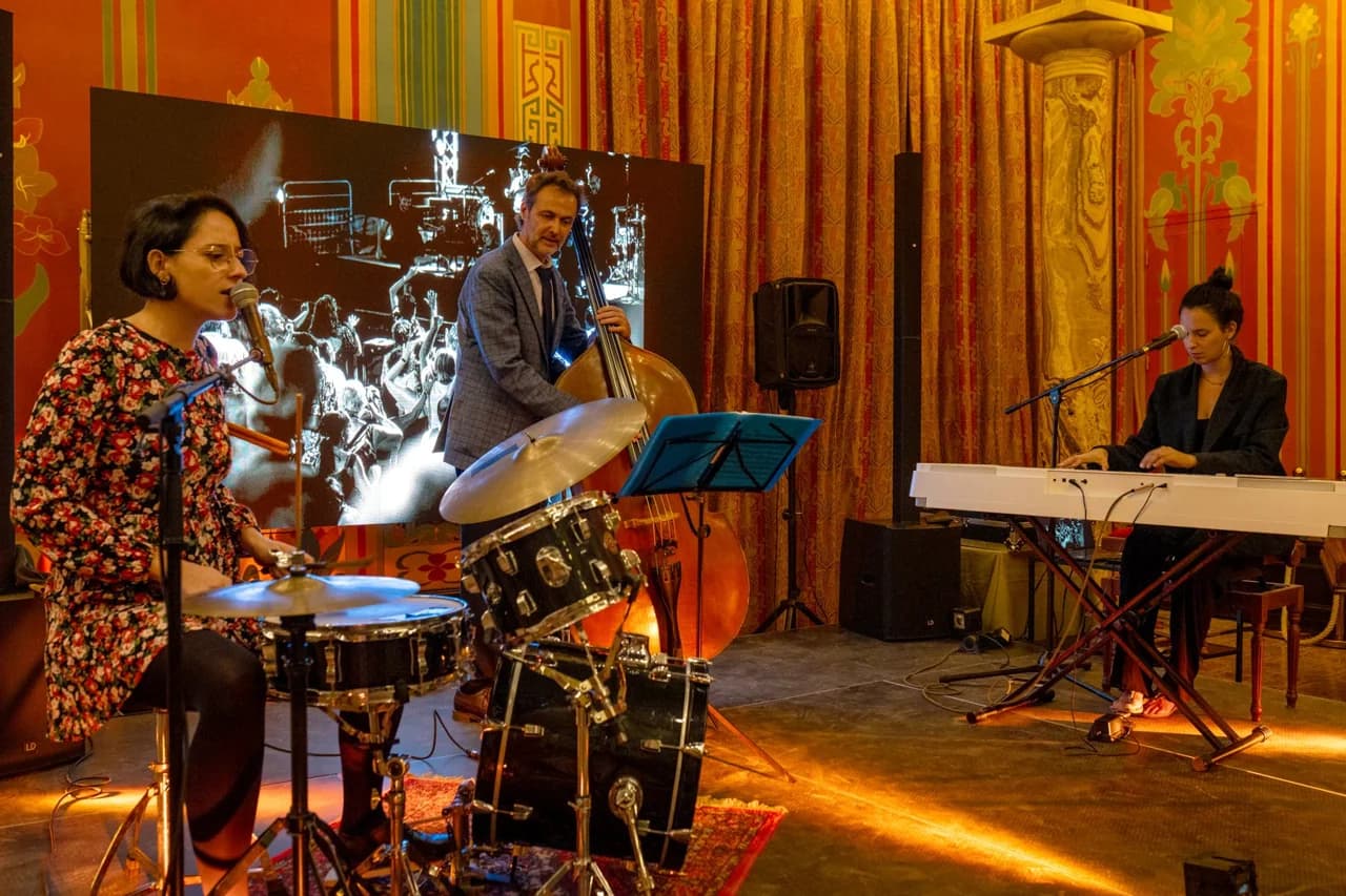 Un trio de sonorité[s], batterie, contrebasse et piano, en concert devant un écran dans une salle aux murs colorés.