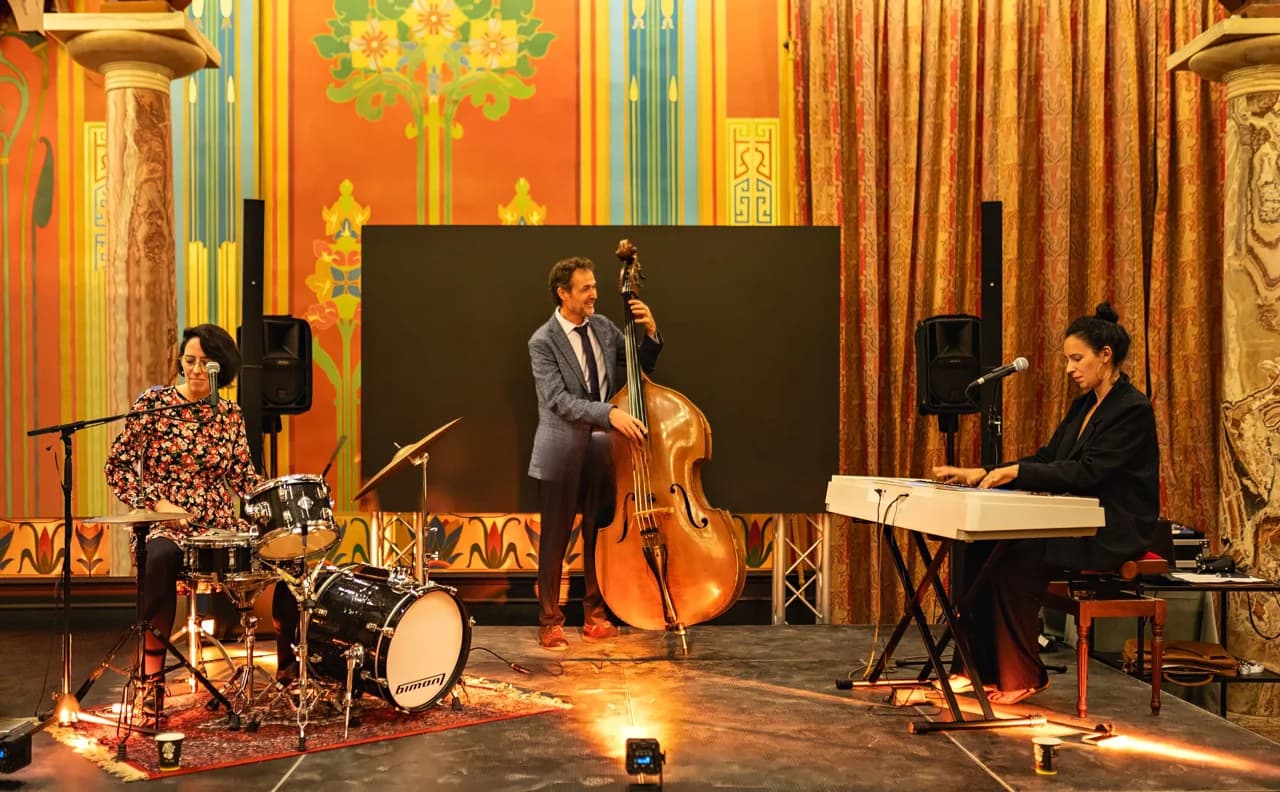 Un trio de sonorité[s] en concert, avec batterie, contrebasse et piano, dans une salle aux boiseries dorées.