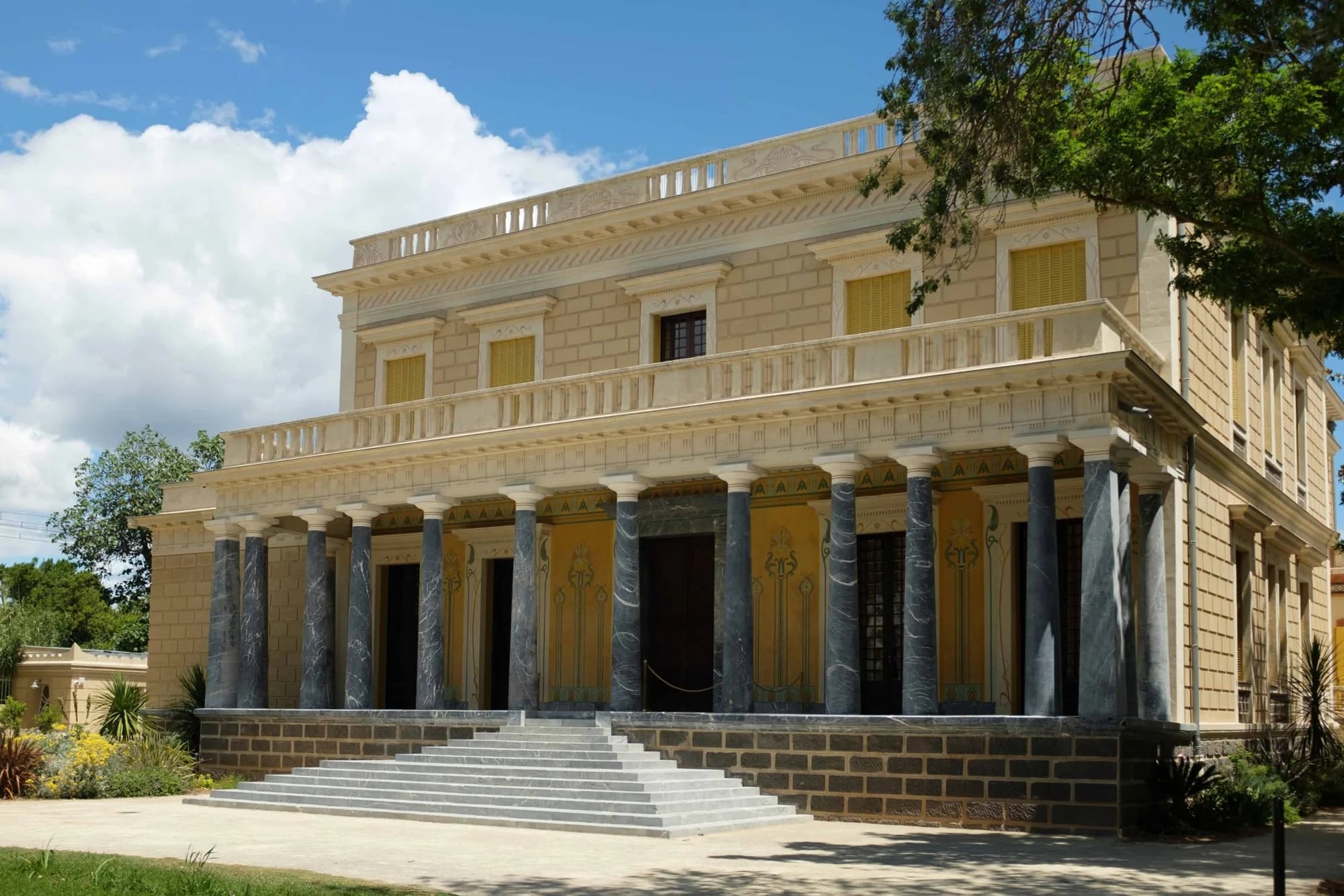 La façade du Château Laurens en plein jour, ses colonnes de marbre et son grand escalier, sous un ciel d’été.