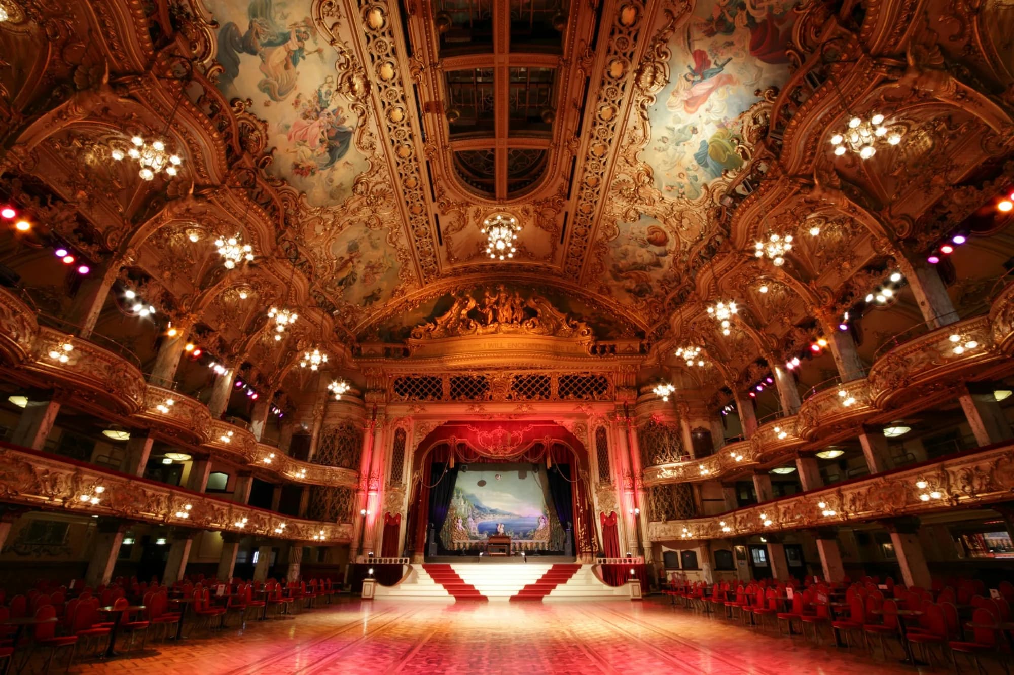 Une grande salle de bal ornée de dorures et de lustres, avec sa scène au rideau rouge, ses balcons et son plafond peint.
