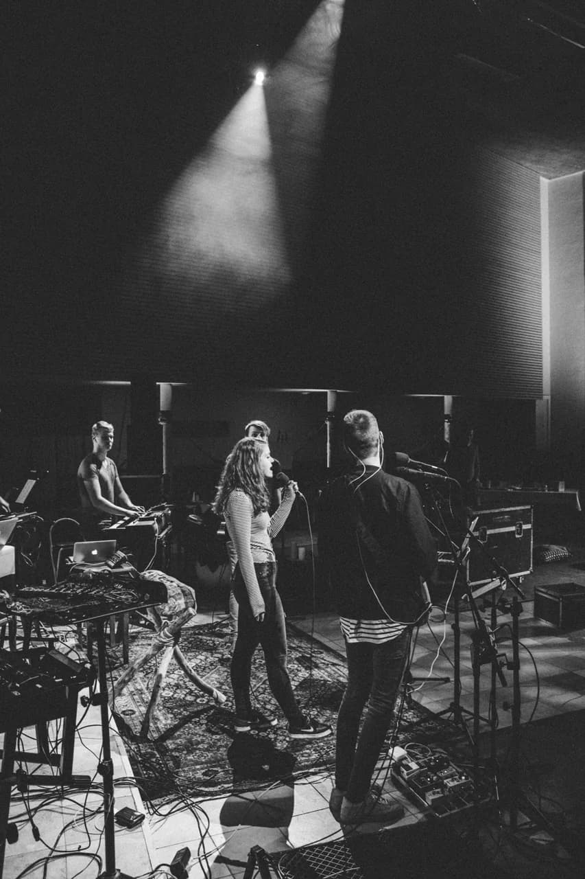 Une chanteuse et des musiciens sur une scène de festival éclairée de projecteurs, en noir et blanc.