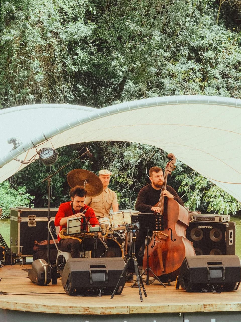Un trio de jazz se produit en plein air, sous une scène couverte entourée d’arbres.