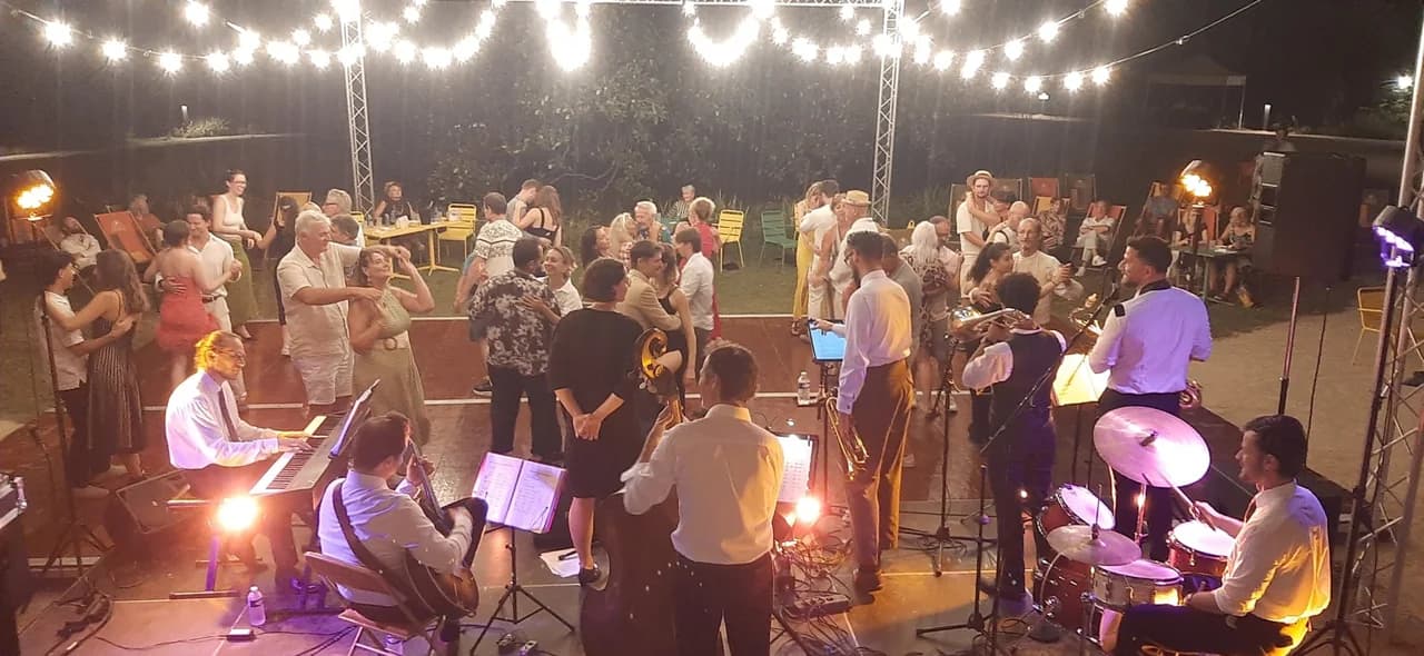 Un grand orchestre de jazz joue en plein air sous des guirlandes lumineuses, devant un public rassemblé.