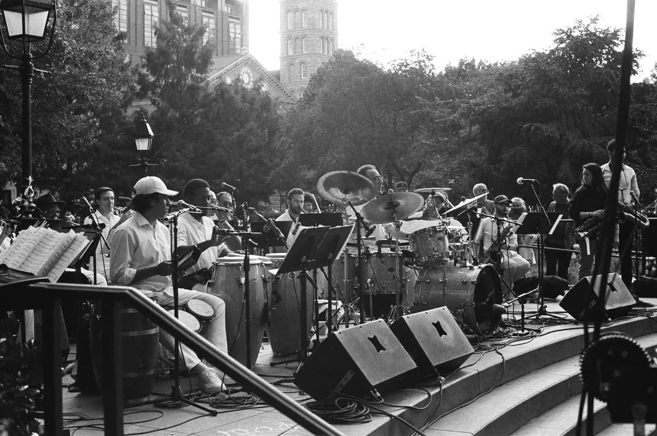 Un big band joue en plein air sur une scène entourée d’arbres, en noir et blanc.