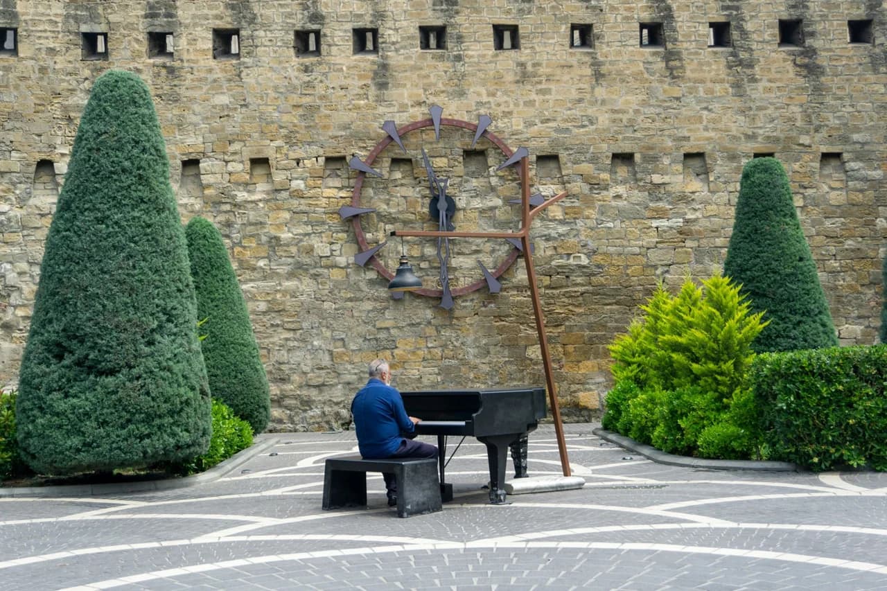 Un pianiste joue sur une place de pierre, devant un mur ancien et deux arbres taillés.