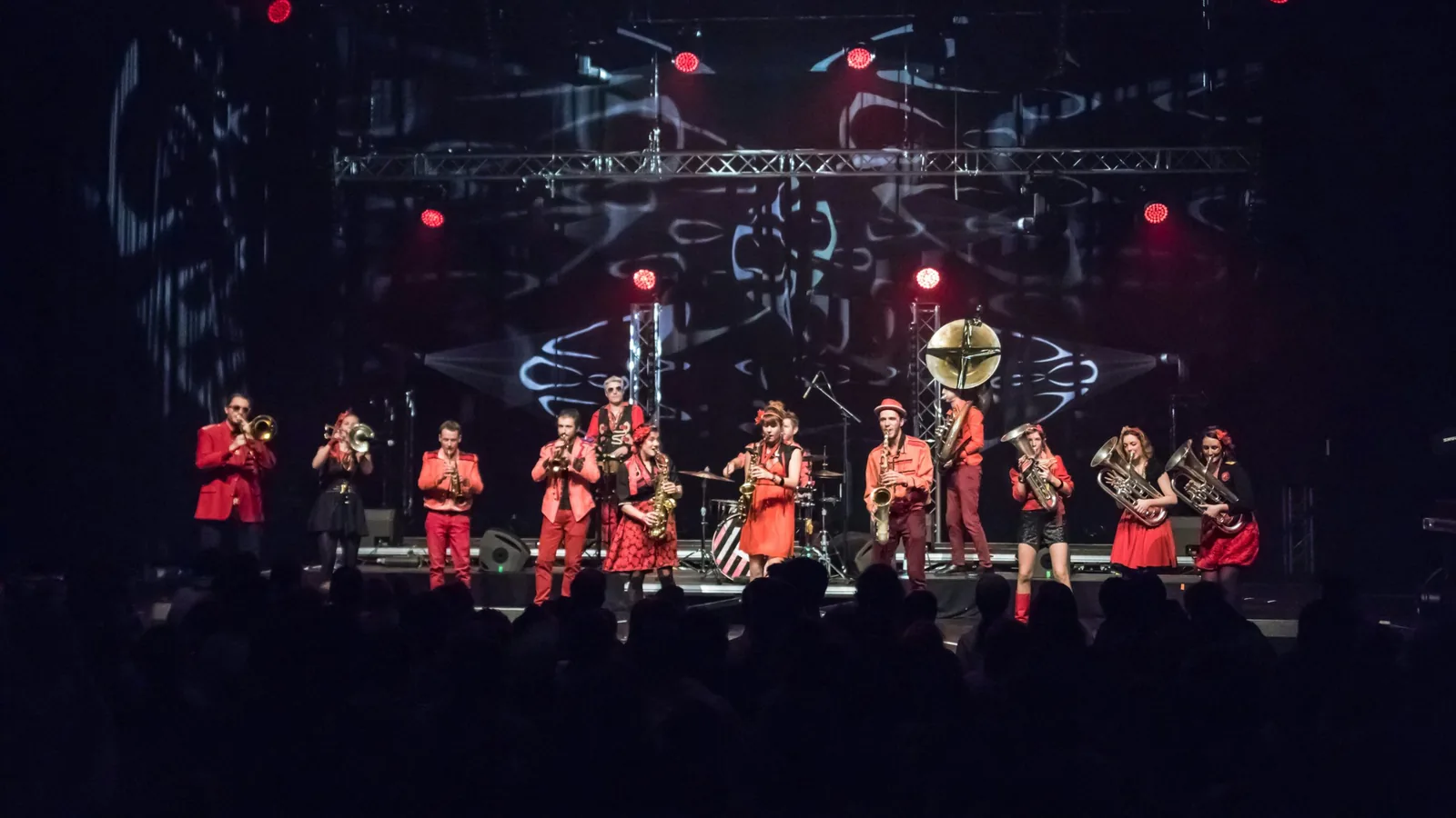 Une fanfare en costumes rouges joue sur une grande scène, cuivres et saxophones en ligne devant le public