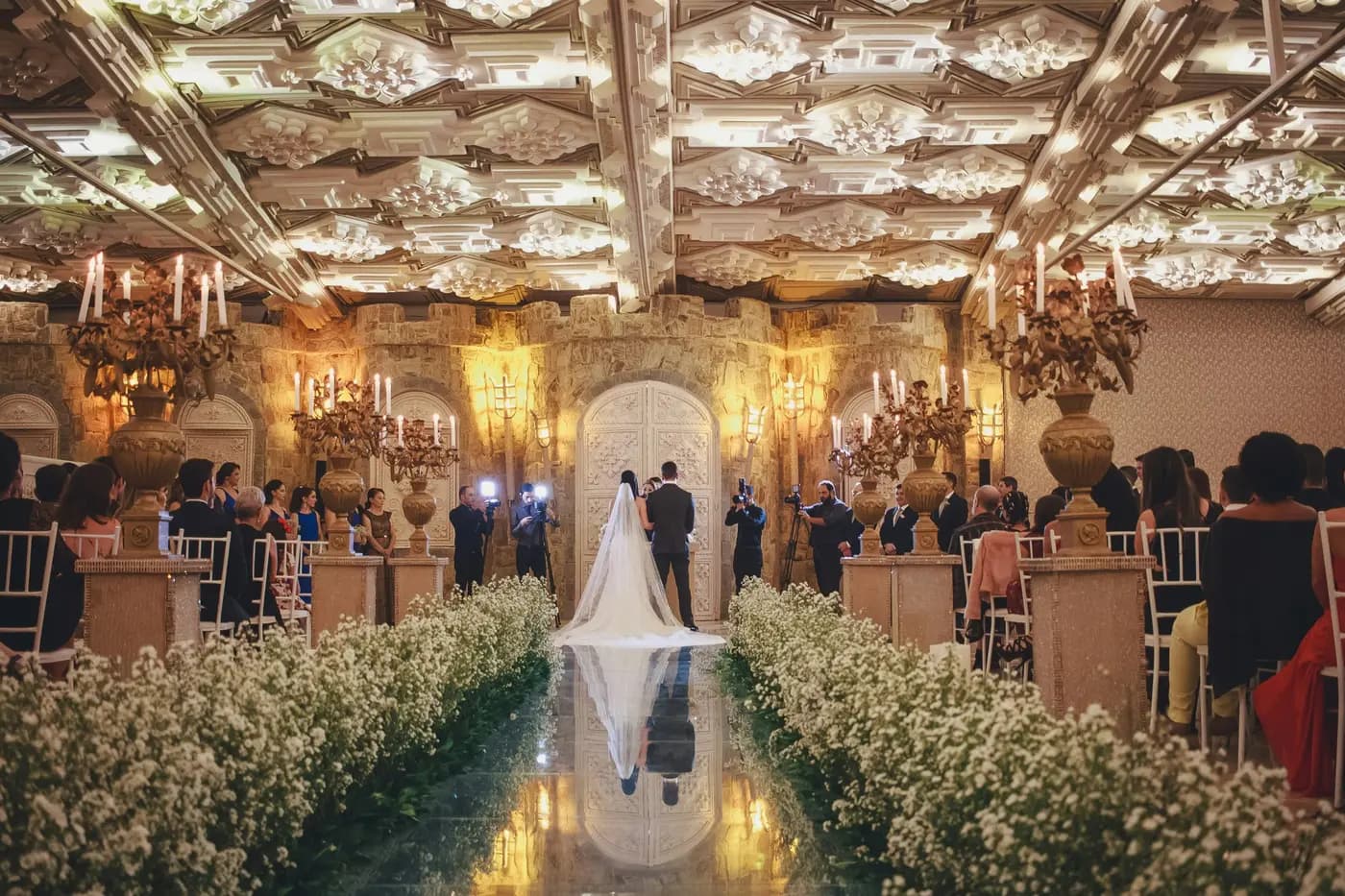 Une cérémonie de mariage dans une salle aux chandeliers, l’allée bordée de fleurs blanches